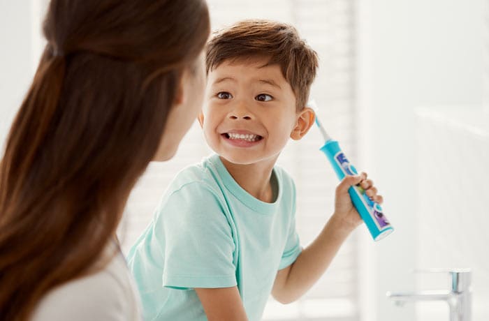 Mały chłopiec szczerzy się do swojej mamy trzymając niebieską szczoteczkę Philips Sonicare for Kids. 