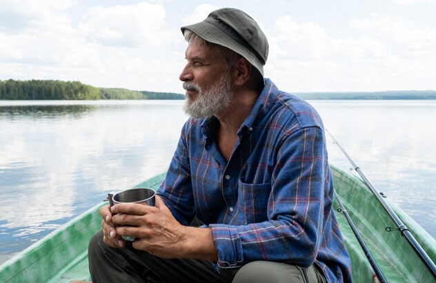 Starszy mężczyzna z siwą brodą, kapeluszem rybackim i niebieską koszulą w kratę siedzi na łodzi i patrzy w dal.