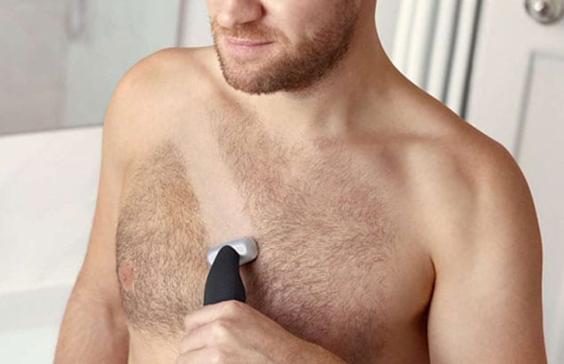 Zbliżenie na owłosioną klatkę piersiową mężczyzny, na której został wygolony poprzeczny pasek za pomocą trzymanej przez niego golarki elektrycznej.