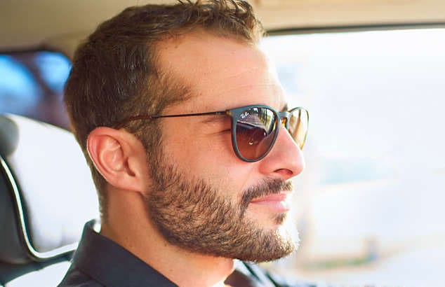 Mężczyzna prowadzący samochód w czarnych okularach przeciwsłonecznych nosi pełną brązową brodę i krótkie wąsy.