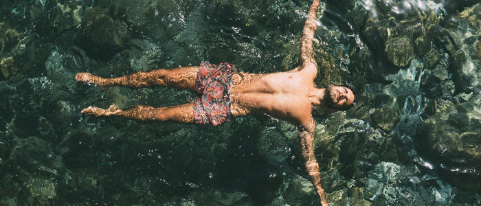 Szeroki kadr z brodatym mężczyzną w szortach kąpielowych pływającym twarzą do góry w wodzie z rozłożonymi ramionami.