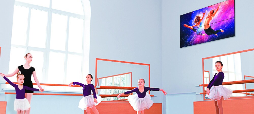 Monitor informacyjno-reklamowy na ścianie. Dzieci uczą się tańczyć balet w pokoju